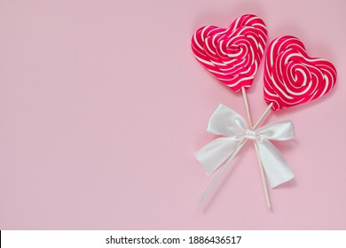 On A Pink Background Are Two Bright Lollipops With A Heart On A Wooden Stick With A White Bow On The Right Side. Greeting Card. Empty Space. Top View. Flatlay Layout. Minimalism.