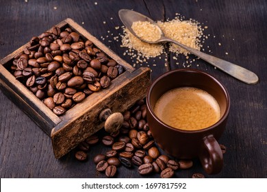 On The Raw And Dark Wooden Table, A Cup Of Coffee With A Spoonful Of Brown Sugar And Roasted Coffee Beans.