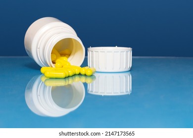 On A Reflective Surface There Is An Overturned Yellow Pill Bottle From Which Some Pills Are Coming Out, Next To It Is The Cap. The Background Is Blue.