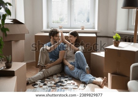 On relocation day happy young wife and husband sit on floor giving high five, boxes with belongings and heap of color samples tiles, renovation brochures nearby. Repair works, renovation, new house