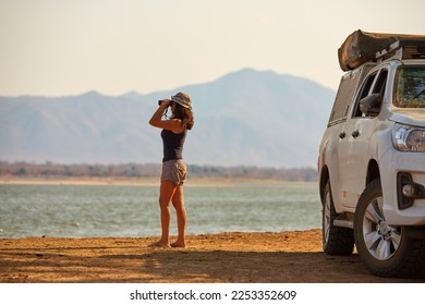 On safari in Zambia: Silhouette of a Fit Woman standing next to white safari car, observing Zambezi River Nature through binoculars. Lower Zambezi National Park, Zambia, Africa.