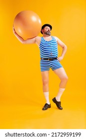 On Sea Beach. Cute Fat Young Man Wearing Retro Striped Swimsuit And Vintage Bowler Hat With Huge Ball Isolated On Bright Yellow Background. Vacation, Summer, Funny Meme Emotions Concept.