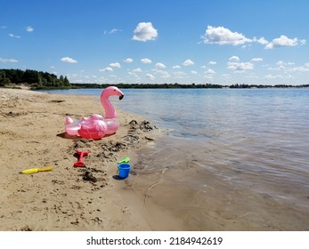 On The Shore Of The Lake Are Decal Toys For The Baby And Inflatable Circle Flamingos. On The Beach There Are Children's Toys And An Inflatable Circle In The Form Of Flamingos.