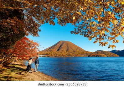 
On the shore of Lake Haruna 榛名湖, a young couple enjoy the view of beautiful fall colors with Mount Haruna-Fuji 榛名富士 under blue clear sky in background on a sunny autumn day, in Takasaki, Gunma, Japan