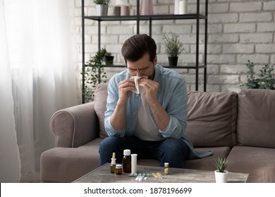 On Sick Leave. Ill Young Man Stay At Home On Workday Sneezing Feeling Bad Catching Cold Flu. Unhealthy Millennial Male Sitting On Sofa Blowing Nose At Paper Tissue Take Pills Medicine For Treatment