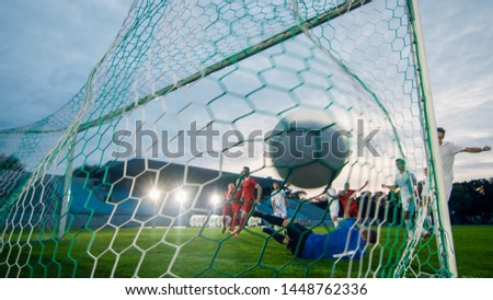On Soccer Championship Goalkeeper Tries to Defend Goals but Jumps and Fails to Catch the Ball. Shot from Behind the Net with the Ball in it. Whole Stadium Visible.