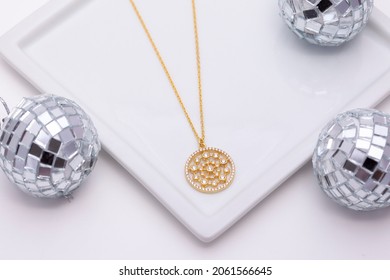 On A Soft White Background, A Gold Necklace On A White Plate And Disco Balls Around It.