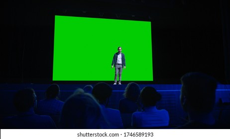 On Stage, Keynote Speaker Presents New Product To The Audience, Behind Him Movie Theater With Green Screen, Mock-up, Chroma Key. Business Live Event Or Device Reveal