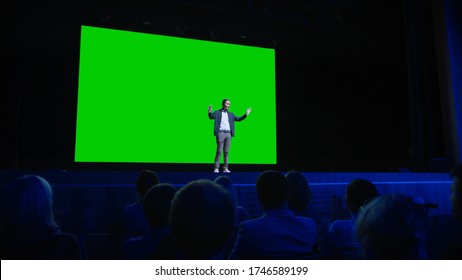 On Stage, Keynote Speaker Presents New Product To The Audience, Behind Him Movie Theater With Green Screen, Mock-up, Chroma Key. Business Live Event Or Device Reveal