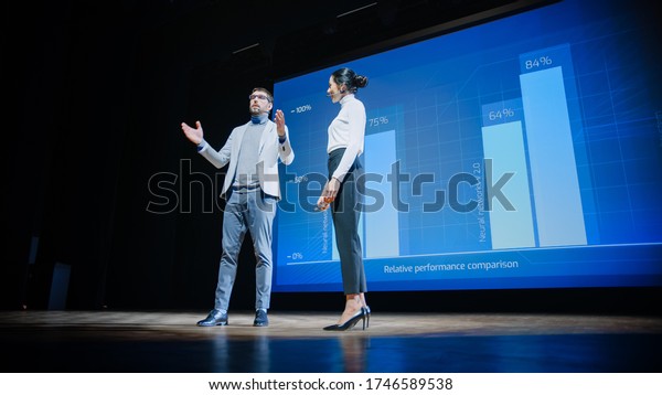 On Stage, Successful Female CEO and Male COO\
Speakers Present Company\'s New Product, Show Infographics,\
Statistics on Big Screen, Talk About Growth. Live Event, Tech\
Startup, Business Conference