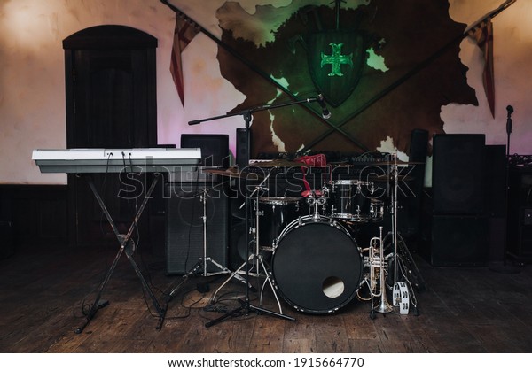 On stage there is a dark drum kit with cymbals\
and a microphone stand, two trumpets (alto, bass), a tambourine and\
a keyboard synthesizer. Spears, shield and sword on the wall.Jazz\
band live concert.