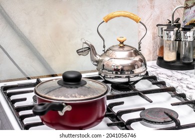 On The Stove There Is A Shiny Ancient Teapot With A Strainer For Brewing Tea, A Burgundy Saucepan With A Handle And Lid, And A Set Of Spices On The Table Next To It