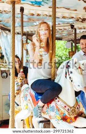 On a summer day a group of friends having fun in the shade on a carousel with horses and blowing soap bubbles. In the foreground a young woman in the air while blowing many colorful soap bubbles