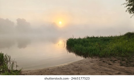 On summer morning, the sun rising over the forest on the far side of the river breaks through the mist billowing over the water. Footprints were imprinted on the sandy beach near the reeds - Powered by Shutterstock