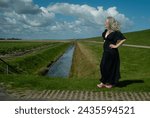 On a summerday a woman in a black long dress en pink wooden shoes is looking forward with dikes, ditch, clouds and a little house in the background and she is satified