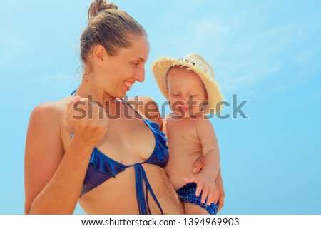 On sunny beach happy young mother hold on hands baby son in straw hat and have fun before swimming in sea. Active children lifestyle, summer vacation travel with kids on tropical family resort.