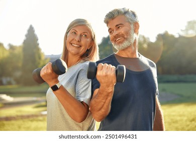 On a sunny day in a vibrant park, a cheerful mature couple lifts dumbbells together, showing their commitment to fitness and healthy aging through fun outdoor exercises accessible to the community - Powered by Shutterstock