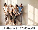 On a sunny morning beautiful diverse girls gathered at gym for workout. Four slim women in sportswear standing barefoot near wall holding yoga mats talking feels happy. Group training wellness concept