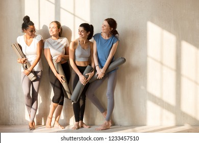 On a sunny morning beautiful diverse girls gathered at gym for workout. Four slim women in sportswear standing barefoot near wall holding yoga mats talking feels happy. Group training wellness concept - Powered by Shutterstock