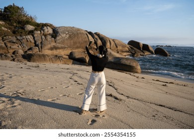 On a sunny morning, a young woman stands barefoot on a sandy beach, using her smartphone to capture the picturesque view of the ocean and rocky coastline - Powered by Shutterstock