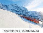 On a sunny winter day, tourists travel on a cogwheel train from Jungfraujoch (Top of Europe) to Kleine Scheidegg on the snowy hillside with Jungfrau in background, in Berner Oberland, Switzerland