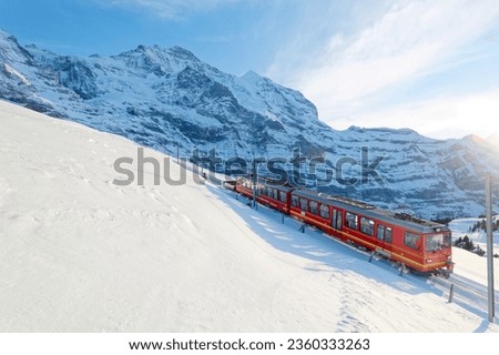 On a sunny winter day, tourists travel on a cogwheel train from Jungfraujoch (Top of Europe) to Kleine Scheidegg on the snowy hillside with Jungfrau in background, in Berner Oberland, Switzerland