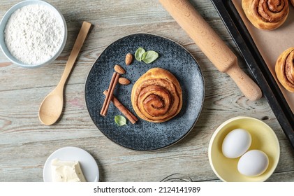 On The Table Is A Baking Tray With Fresh Cinnamon Rolls, Ingredients For Their Cooking And An Appetizing Bun On A Plate. Concept Of Tasty Homemade Pastries And A Cozy Atmosphere. Top View.