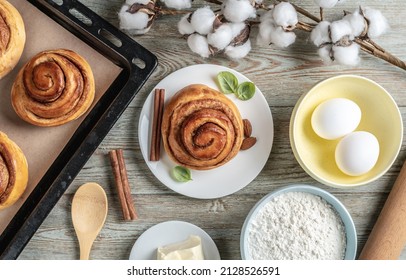 On The Table Is A Baking Tray With Fresh Cinnamon Rolls, Ingredients For Their Cooking And An Appetizing Bun On A Plate. Concept Of Tasty Homemade Pastries And A Cozy Atmosphere. Top View.