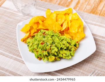 On table in cafe there is plate with traditional Mexican snack guacamole and pile of crispy potato chips. Salty and savory light repast for beer