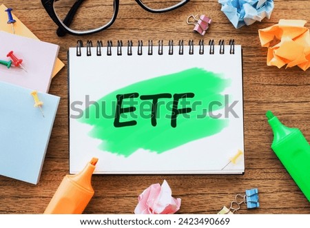 On the table are glasses, coloured markers, crumpled sheets, paper clips and a white notepad with text ETF Exchange Traded Funds highlighted in green. Business concept
