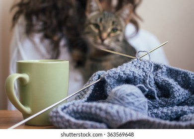 On the table is a knitting and a cup. In the background a girl and a cat.