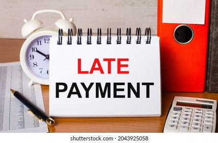 On the table are reports, a white alarm clock, a calculator, folders for papers, a pen and a white notebook with the LATE PAYMENT. Business concept - Shutterstock ID 2043925058