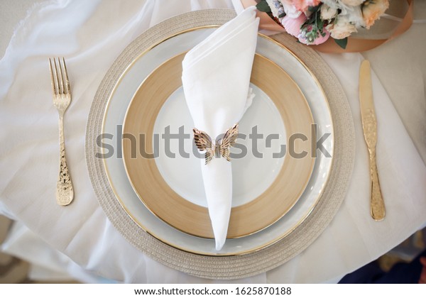 On the table is a set\
of plates with a gold border. On a plate lies a napkin with a\
serving ring in the shape of a butterfly. On the left is a fork, on\
the right is a knife.