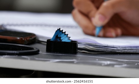 On a table, there is a pencil sharpener filled with pencil shavings captured in a detailed close-up. In the blurred background, a hand is visible writing something in a notebook with a pencil - Powered by Shutterstock