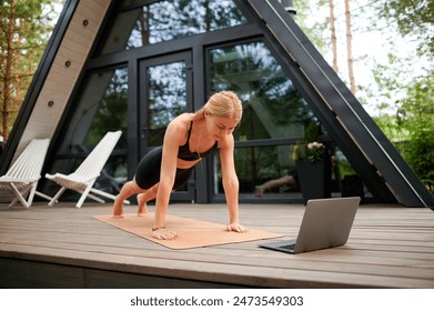 On the terrace of her house in the summer, a young pretty European girl engages in a workout with a video call coach, emphasizing virtual fitness, outdoor exercise, healthy habits, summer training - Powered by Shutterstock