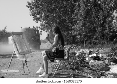 
On This Black And White Photo, A Beautiful Girl With A Long, Lush Hair Is Sitting On The Street In A Chair And Painting. In The Distance, You Can See The Truck Passing Through And Raising Dust