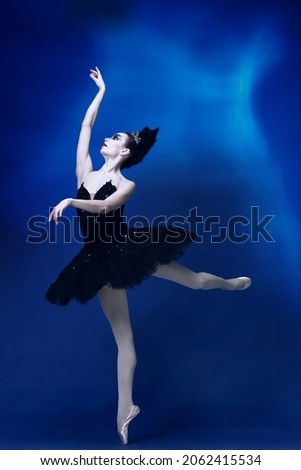 On tips of toes. Portrait of young beautiful woman, ballerina in black ballet outfit, tutu dancing at blue studio full of light. Classical ballet, famous performance, art concept. Passion in dance