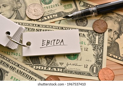 On top of the dollar bills on the table, there is a word book with the financial term EBITDA written on it. It is an abbreviation for Earnings Before Interest Taxes Depreciation and Amortization.
