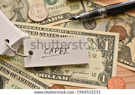 On top of the dollar bills on the table, there is a word book with the financial term CAPEX written on it. It is an abbreviation for Capital expenditure.