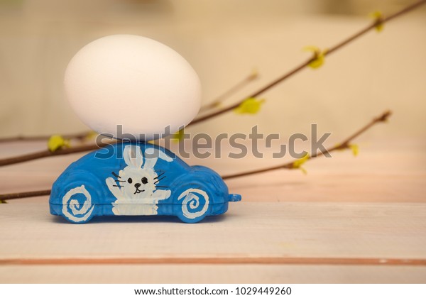 On a toy car there is a chicken egg.\
On the door of the car the Easter rabbit is depicted. Easter\
motive. In the background a branch with young green\
leaves