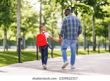 On way to school. Ethnic son schoolboy with backpack holding hand of father, boy looking at dad with smile while going to first grade on sunny autumn day through park  - Powered by Shutterstock