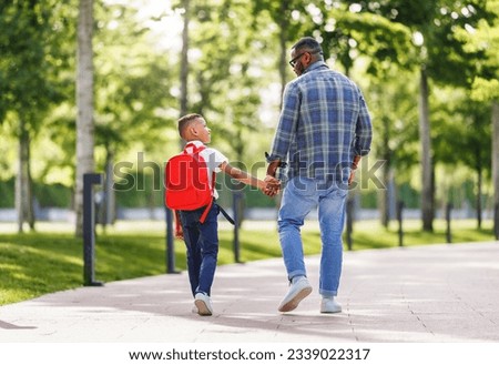 On way to school. Ethnic son schoolboy with backpack holding hand of father, boy looking at dad with smile while going to first grade on sunny autumn day through park 