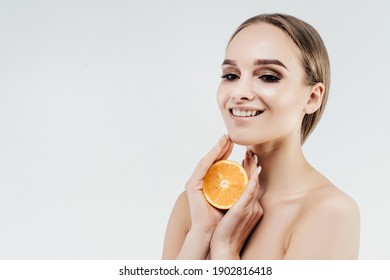 On A White Background, A Pretty Woman With Radiant Skin Holding Half An Orange Near Her Face And Smiling. Health And Beauty. Beautician.