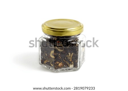 On a white background there is one glass jar of black tea with dry leaves and dried fruits.