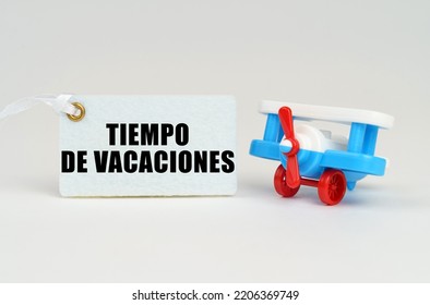 On A White Surface There Is A Toy Airplane And A Sign With The Inscription - VACATION TIME. Text In Spanish.