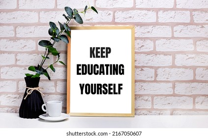 On A White Table Against A Light Brick Wall, A Branch Of Eucalyptus In A Dark Vase, A White Cup And A Light Wooden Frame With The Text KEEP EDUCATING YOURSELF. Home Office Interior.