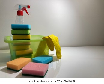 On The White Table There Is A Cleaning Kit For The Apartment. Green Plastic Basin, Spray Bottle With Detergent, Yellow Rubber Gloves, And Lots Of Sponges In Different Colors.