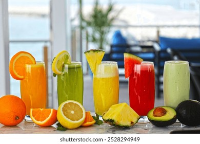 On a white table there are various types of juice such as orange, lemon, pineapple, watermelon and avocado, with a blue chair and white window in the background - Powered by Shutterstock