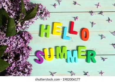 on a wooden background there is an inscription in multicolored letters hello summer surrounded by lilac flowers