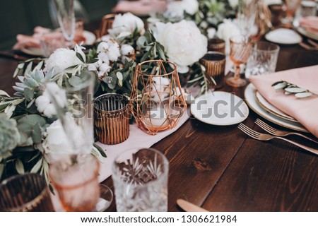 on wooden banquet table are glasses, plates, candles, table is decorated with compositions of cotton and eucalyptus branches, plates are decorated with napkins and sprig of Italian greenery